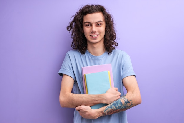 Funny young caucasian guy with books notebooks in casual tshirt posing isolated on purple wall backg...