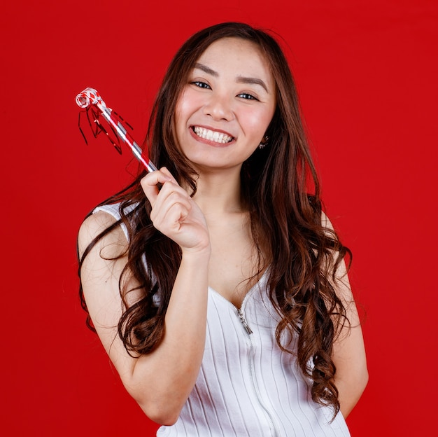 Funny young and cure Asian girl holding and playing party whistle with funny and happy. Studio shot on red background.