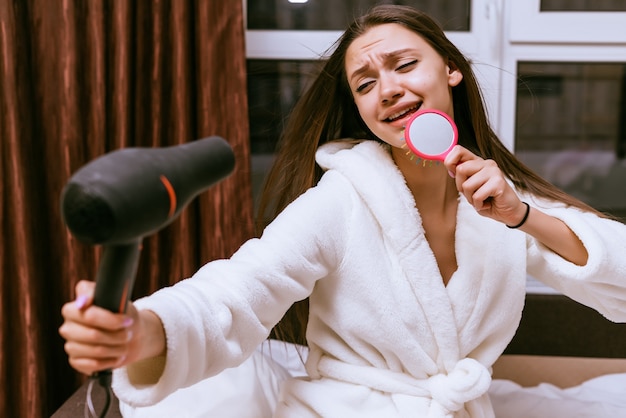 Funny young girl dries her long hair with a hair dryer, sings in a comb