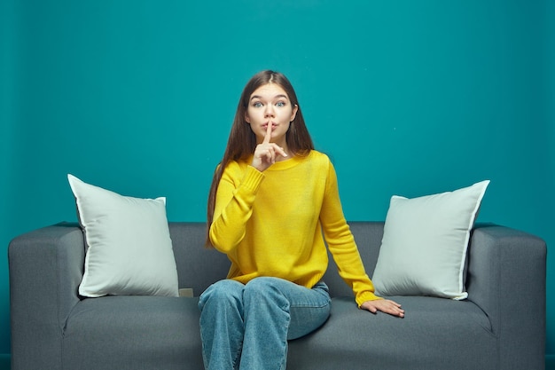 Funny young girl show silence gesture ask to be quiet sitting on sofa watching modern interesting