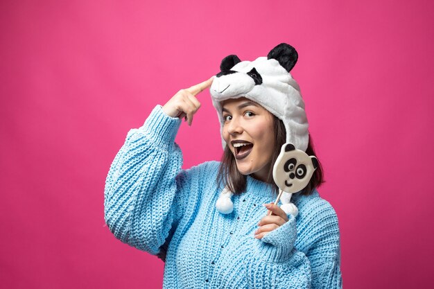 Photo funny young girl standing with yummy pandalollipop in her hand and a hat on his head on a pink