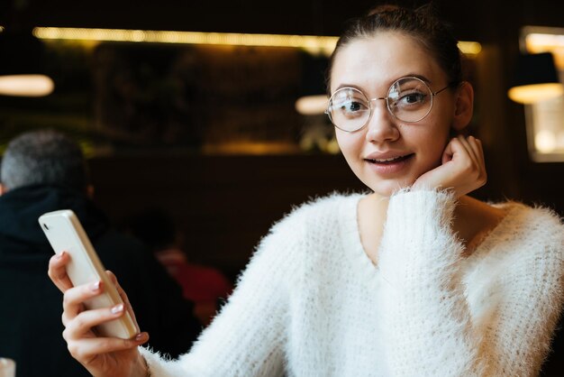 Funny young girl student wearing glasses looks surprised holds smartphone in cafe