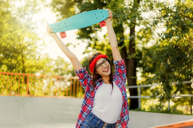 Funny young hipster woman dressed in stylish clothes holds skateboard in her hand in skatepark at bright sunny day