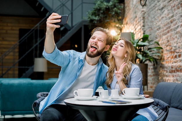 Funny young loving couple making selfie photo on the smartphone together while sitting in cafe or hotel and having coffee