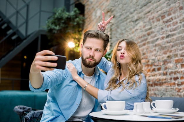 Funny young loving couple making selfie photo on the smartphone together while sitting in cafe or hotel and having coffee