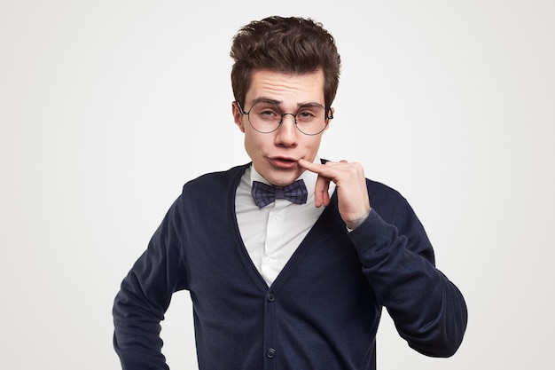 Funny young male student in elegant suit and nerdy glasses touching lips and looking at camera against white background