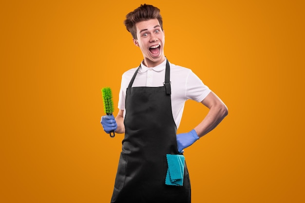 Funny young man in black apron and rubber gloves with napkin and brush looking at camera with astonished face expression, while doing household chores against yellow background
