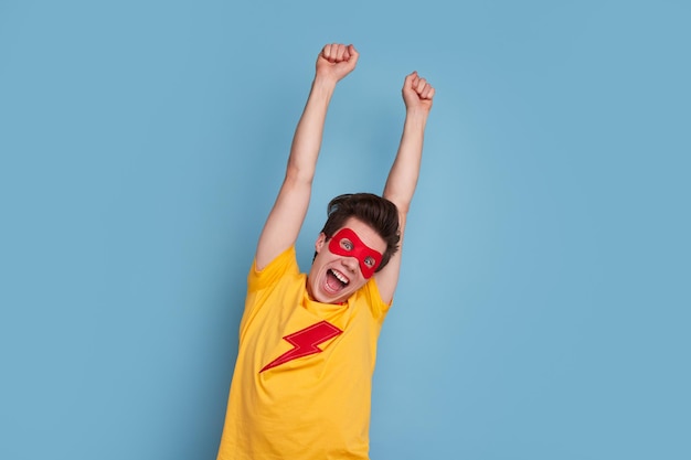 Funny young man in colorful superhero t shirt and mask raising arms and yelling in amazement against blue background