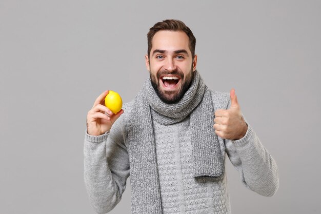 Funny young man in gray sweater, scarf posing isolated on grey
background in studio. healthy fashion lifestyle, people emotions,
cold season concept. mock up copy space. hold lemon, showing thumb
up.