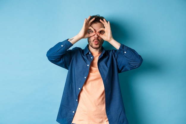 Funny young man looking through hand binoculars and say wow, checking out something interesting, standing amazed on blue background.