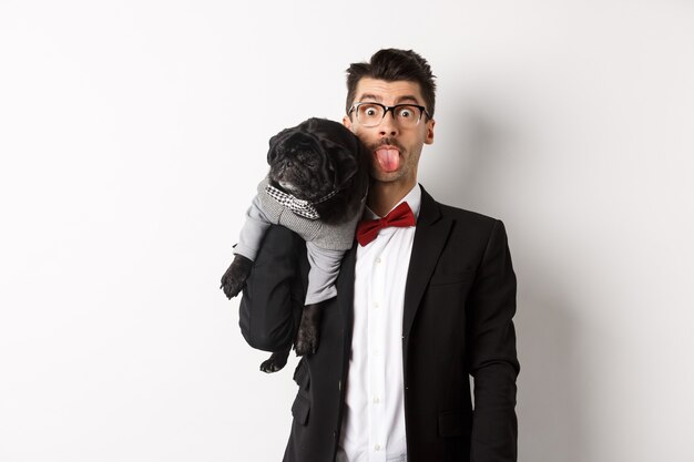 Funny young man in party suit, showing tongue and holding cute black pug on shoulder, celebrating with pet, standing over white.