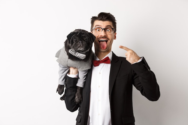 Funny young man in party suit, showing tongue and making grimaces, pointing at cute black dog in winter clothes, standing over white.