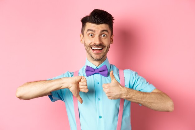 Funny young man smiling and showing thumbs up down, judging something and being indecisive, making choice, standing on pink.