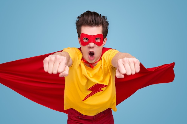 Funny young man in superhero costume roaring and flying towards camera while saving world against blue background