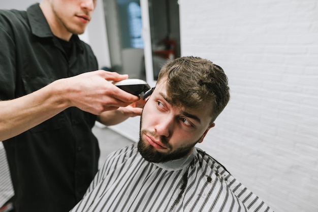 Забавный молодой человек с бородой стрижет парикмахера с недовольным лицом, смотрит в сторону