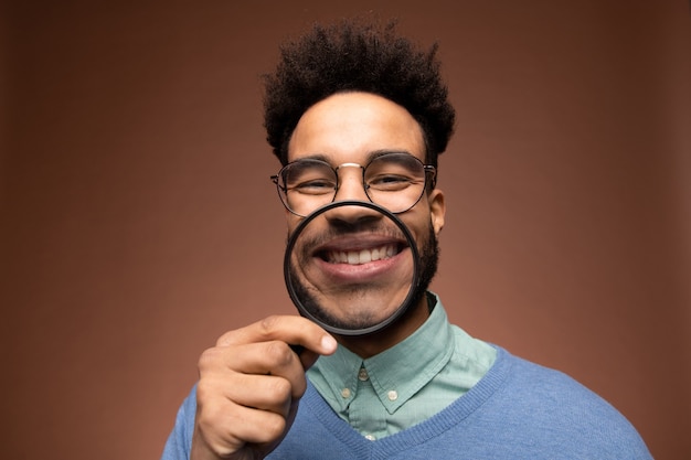 Funny young mixed-race businessman in smart casualwear looking at you while holding magnifying glass in front of his toothy smile