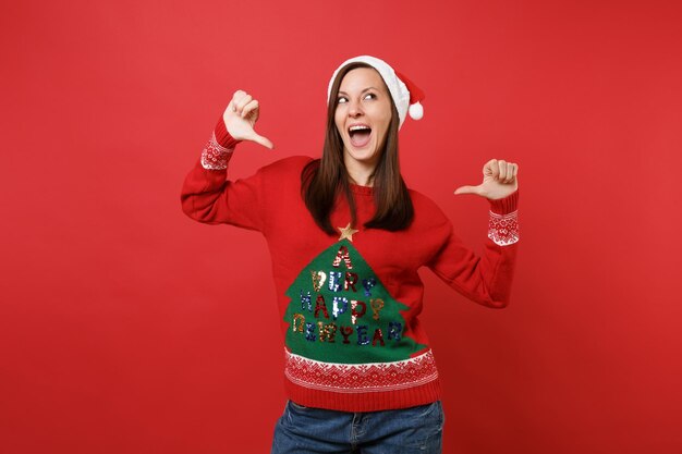 Funny young Santa girl in Christmas hat looking up keeping mouth open, pointing thumbs on herself isolated on red background. Happy New Year 2019 celebration holiday party concept. Mock up copy space.