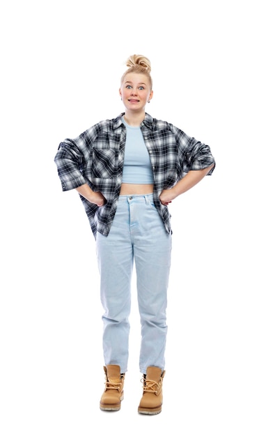 A funny young teenage girl stands with her hands on her belt Laughing blonde girl in jeans blue top and plaid shirt Isolated on a white background Vertical Full height