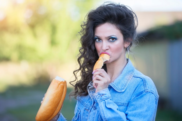 Photo funny young woman in a denim jacket with make-up in the style of the eighties eats a loaf.