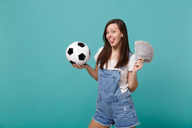 Funny young woman football fan support favorite team with soccer ball, fan of money in dollar banknotes, cash money, showing tongue isolated on blue turquoise background. Sport family leisure concept.