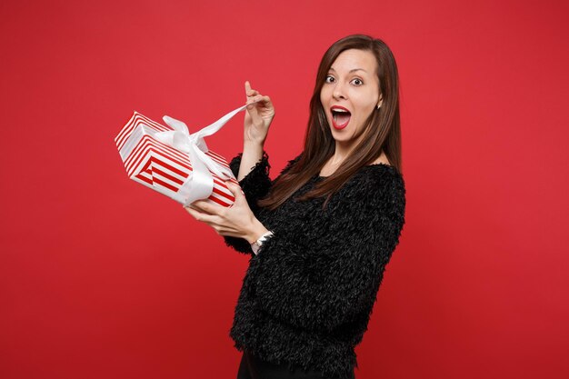 Funny young woman in fur sweater opening red striped present box with gift ribbon isolated on red background. Valentine's Day, International Women's Day, birthday, holiday concept. Mock up copy space.