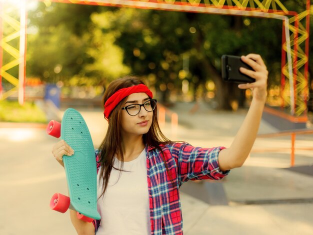 Funny young woman in glasses and red plaid shirt. Hipster girl n skatepark while makes  selfie with skateboard in skatepark at sunny bright day.  Summer fun