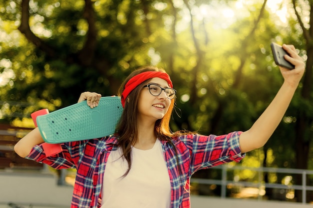 안경 및 붉은 격자 무늬 셔츠에 재미있는 젊은 여자. 소식통 소녀 n skatepark는 화창한 밝은 날에 skatepark에서 스케이트 보드로 셀카를 만드는 동안. 여름의 즐거움