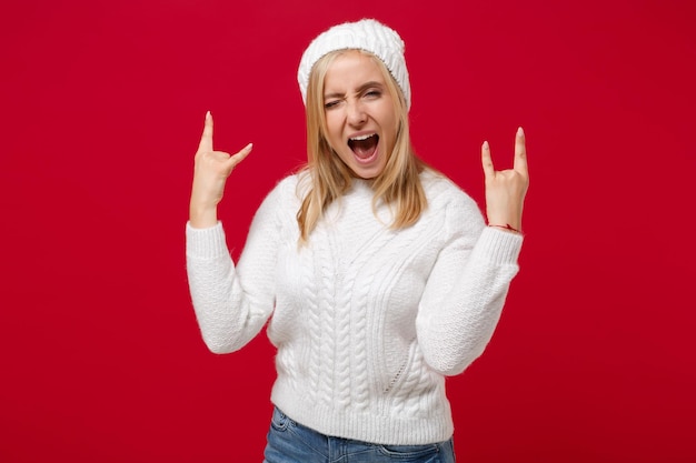 写真 白いセーターを着たおかしな若い女性、赤い壁の背景に隔離された帽子。健康的なファッション ライフ スタイル、寒い季節のコンセプト。コピー スペースをモックアップします。ヘヴィメタルロックのサインを描いた、角を上げるジェスチャーを示す。