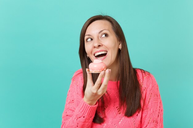 Funny young woman in knitted pink sweater looking up, holding in hand cake isolated on blue turquoise wall background, studio portrait. People sincere emotions, lifestyle concept. Mock up copy space.