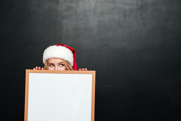 Foto divertente giovane donna con cappello di babbo natale che si nasconde dietro una lavagna bianca vuota blank