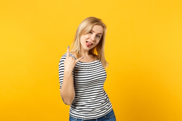 Funny young woman in striped clothes, showing tongue, depicting heavy metal rock sign, rock-n-roll isolated on yellow orange background. People sincere emotions, lifestyle concept. Mock up copy space.