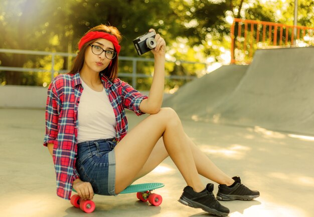 세련 된 옷을 입고 재미있는 젊은 여자는 스케이트 보드에 앉아서 밝고 화창한 날에 skatepark에서 레트로 카메라를 사용합니다. 청소년 개념