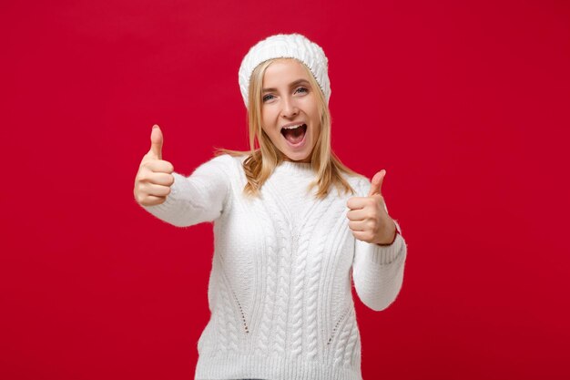 Foto divertente giovane donna in maglione bianco, cappello isolato su sfondo muro rosso, ritratto in studio. stile di vita di moda sano persone emozioni sincere concetto di stagione fredda. simula lo spazio della copia. mostrando i pollici in su.