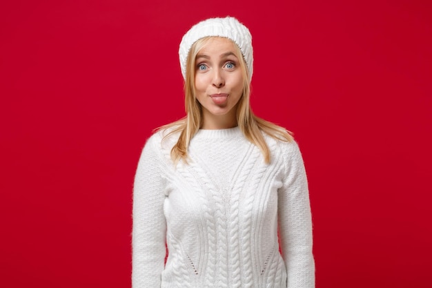 Foto divertente giovane donna in maglione bianco, cappello isolato su sfondo muro rosso, ritratto in studio. stile di vita sano della moda, emozioni sincere della gente, concetto di stagione fredda. simula lo spazio della copia. mostrare la lingua.