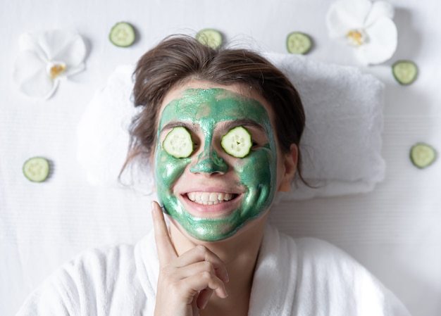 Funny young woman with a cosmetic mask on her face and cucumbers on her eyes