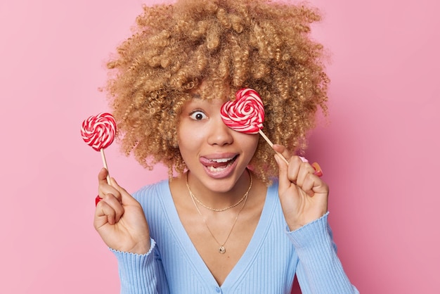 Фото Забавная молодая женщина с вьющимися густыми волосами держит сердечные конфеты на палочках, имеет сахарную зависимость, высовывает язык, носит случайный синий джемпер, изолированный на розовом фоне веселая женщина с леденцом на палочке