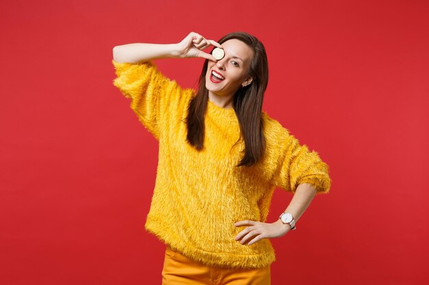 Divertente giovane donna in maglione di pelliccia gialla che copre l'occhio con bitcoin, valuta futura isolata su sfondo rosso brillante della parete in studio. persone sincere emozioni, concetto di stile di vita. mock up copia spazio.