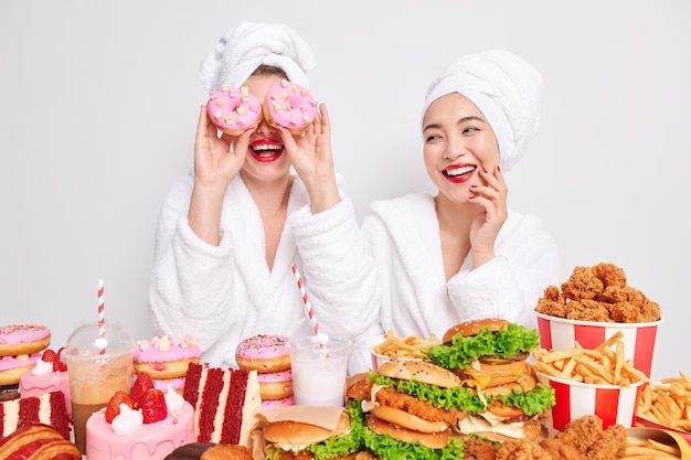 Веселые молодые женщины проводят свободное время дома, глупо храня вкусные сладкие пончики над глазами в окружении вкусного фаст-фуда