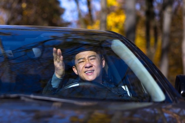 Furious angry asian man standing in a traffic jam beeps and hurries. Sitting in the car. Irritated young business male tired driver in automobile. Stressful commuting to be late work