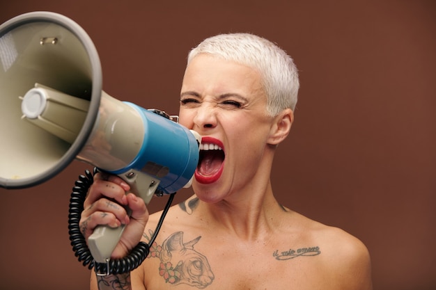 Furious and annoyed blond manlike female with tattoos on arm, chest and neck shouting in megaphone while making important announcement