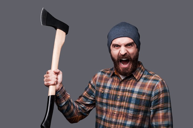 Furious lumberjack. Furious young bearded man swinging his axe and shouting while standing against grey background