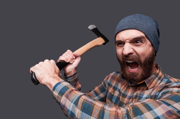 Furious lumberjack. Furious young bearded man swinging his axe and shouting while standing against grey background