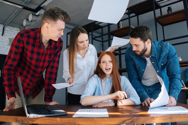 Furious sad young business woman under stress throwing\
documents sitting at desk looking at camera, multi-ethnic\
colleagues standing near her. tired female boss exhausted by\
management work at office.