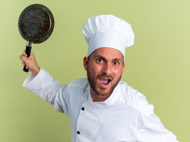 Furious young caucasian male cook in chef uniform and cap raising frying pan looking at camera screaming isolated on olive green wall