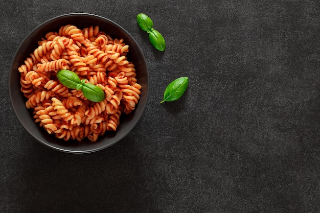 Fusilli pasta cooked with tomato sauce and basil in bowl on dark background top view space to copy text