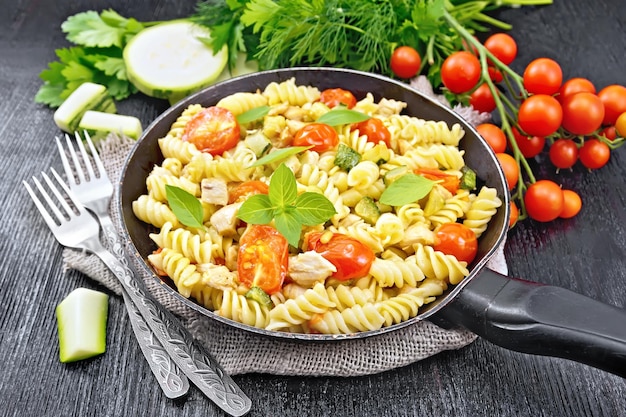 Fusilli with chicken, zucchini and tomatoes in a frying pan on burlap, forks, basil and parsley on wooden board background