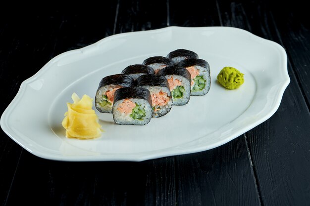 Futomak sushi roll with salmon, cucumber in a white plate on a black wooden background with ginger and wasabi.