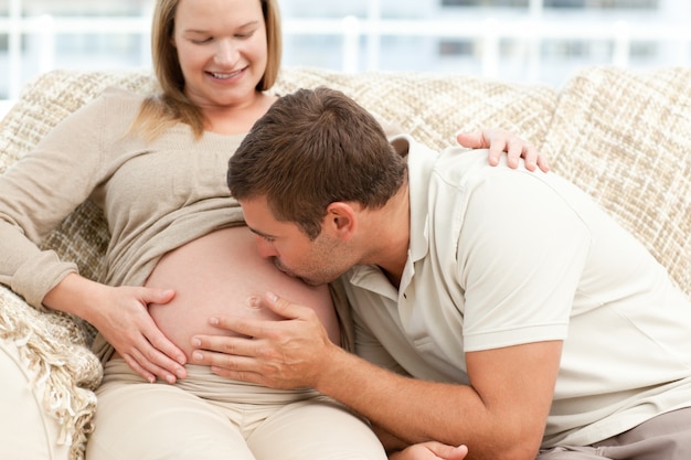 Future dad kissing the belly of his wife sitting on the sofa