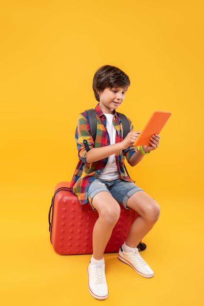 Future explorer. Happy lovely boy using his tablet while sitting on his luggage