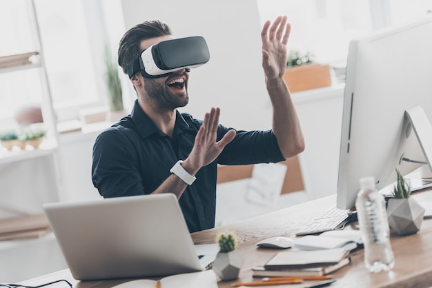 Future is now. Handsome young man in VR headset gesturing and smiling while sitting in creative office